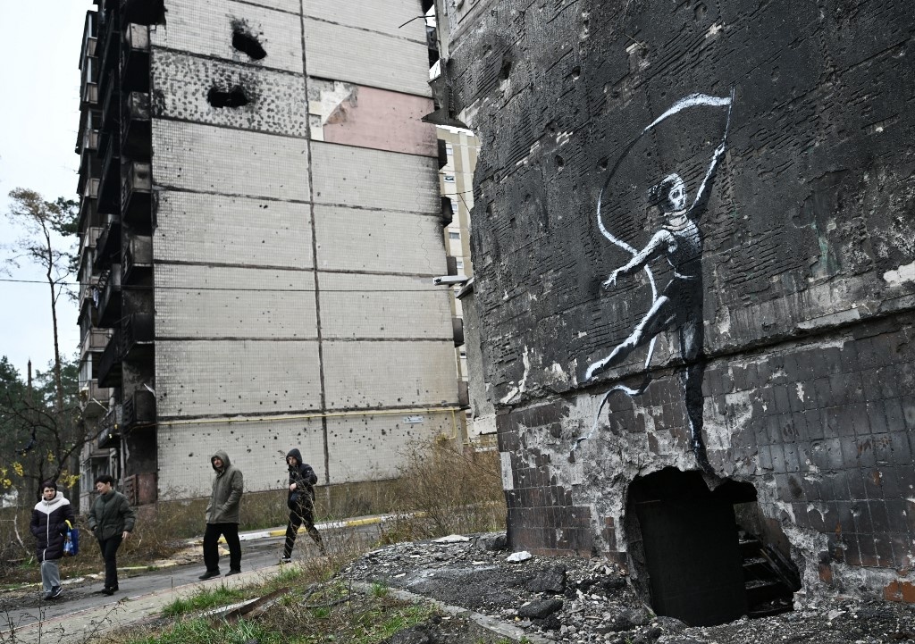 Local residents walk past a graffiti on the wall of a destroyed residential building in Irpin.