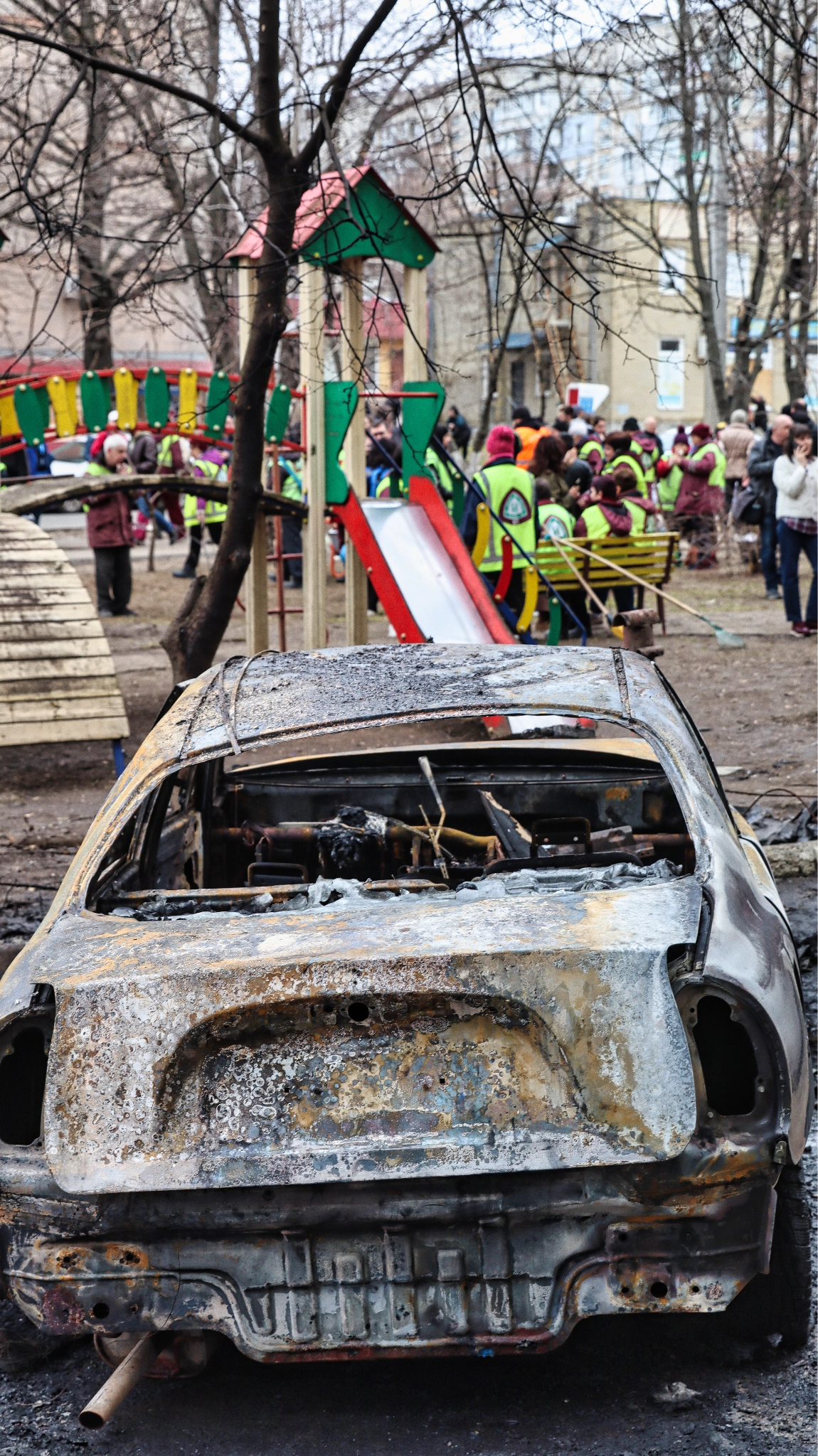 A burned-out car beside a playground in Kharkiv’s Kholodnohirskyi district after a Russian Shahed drone struck the residential neighborhood. Residents and municipal workers gathered within hours to begin clearing the area. April 2026 (Photo by Korbinian Leo Kramer)